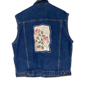 Könnte beinhalten: Eine ärmellose blaue Jeansweste mit einem dekorativen Aufnäher auf dem Rücken. Der Aufnäher zeigt ein Blumendesign mit roten Rosen und grünen Blättern. Die Weste hat einen Knopfverschluss und einen Kragen.
