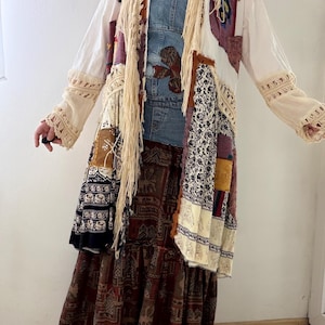 Könnte beinhalten: Ein Patchwork-Mantel mit einem Jeans-Oberteil und einem langen, gestuften Rock. Der Mantel weist verschiedene Stoffmuster auf, darunter Blumen- und geometrische Designs, mit cremefarbenen Fransen und Spitzentrimm. Der Rock hat ein braunes und rotes Muster.