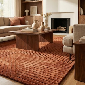 May include: A large, rectangular area rug in a deep terracotta orange colour, featuring a textured surface with raised, parallel linear patterns arranged in geometric sections that create a subtle, repeating grid design across the floor.