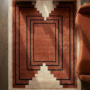 May include: A rectangular area rug with a geometric design. The rug features a central terracotta-colored field framed by black lines and a cream-colored border. Two armchairs and a sofa are visible.