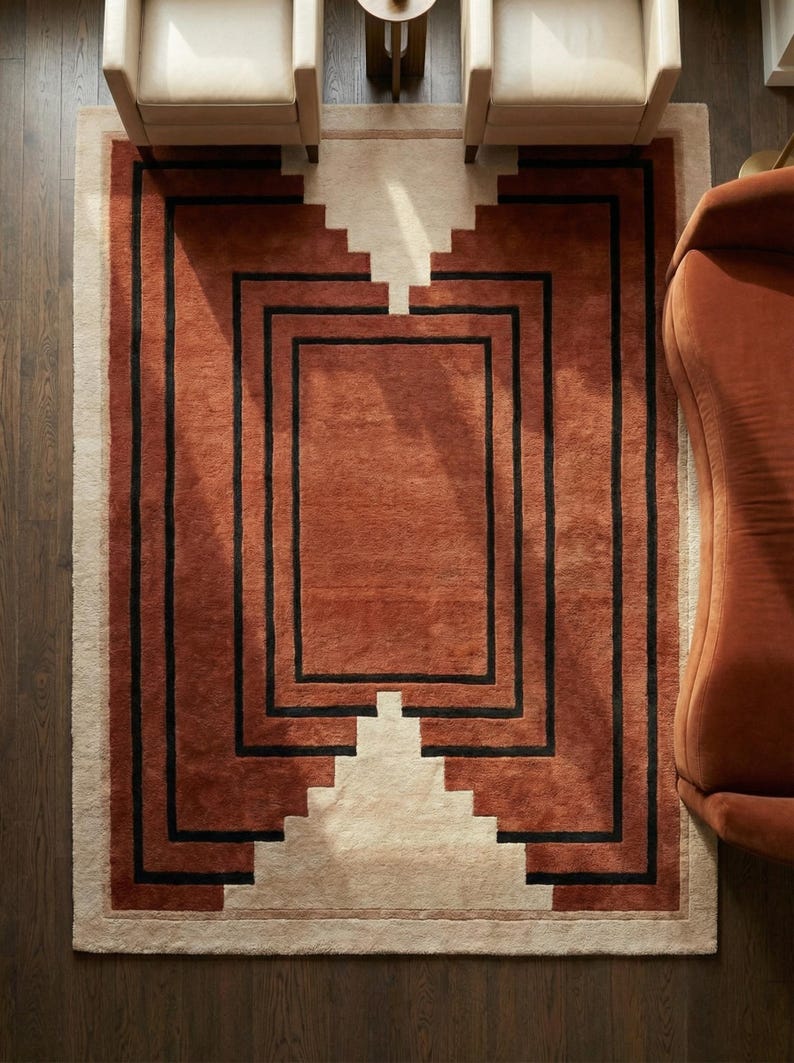 May include: A rectangular area rug with a geometric design. The rug features a central terracotta-colored field framed by black lines and a cream-colored border. Two armchairs and a sofa are visible.