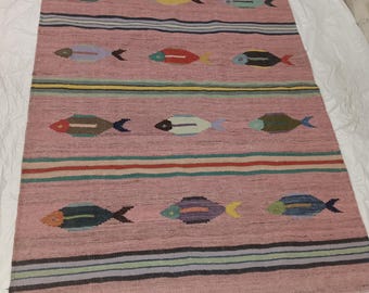 May include: A rectangular pink rug with a repeating pattern of colorful fish. The fish are in various colors, including red, yellow, and blue. Horizontal stripes in multiple colors separate the rows of fish. The rug is made of woven material.