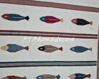 May include: Cream-colored woven rug with a repeating pattern of colorful fish. The fish are in shades of blue, brown, red, and green, with a striped border. The rug has a fringed edge and the text "ATAhomedecore".