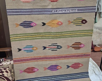 May include: Beige rectangular rug with a repeating pattern of colorful fish. The fish are striped in various colors, including pink, blue, and yellow. Horizontal stripes in blue, green, and pink separate the rows of fish. The text "ATAhomedecore" is visible.