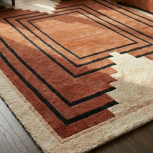 May include: A rectangular area rug with a geometric design. The rug features a central section in a warm, reddish-brown hue, framed by black lines. The outer edges transition to a lighter beige color, creating a layered effect. The rug is placed on a dark wood floor.
