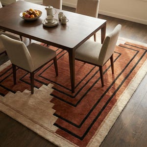 May include: A dining room scene featuring a dark wood table with four beige chairs. A patterned rug in shades of brown and beige with black accents is beneath the table. A bowl of fruit and a tea set are on the table.