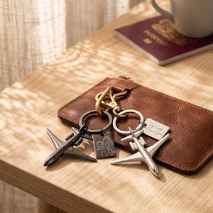 Könnte beinhalten: Eine Sammlung von Flugzeug-Schlüsselanhängern und eine braune Ledergeldbörse auf einem Holztisch. Ein Schlüsselanhänger trägt den Text "Fly Safe I Love You". Ein Reisepass und eine Tasse befinden sich im Hintergrund.