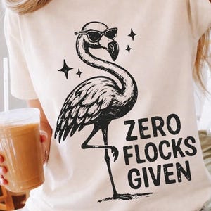 Puede incluir: Camiseta color crema con una ilustración negra de un flamenco con gafas de sol y el texto "ZERO FLOCKS GIVEN". El flamenco está rodeado de estrellas. Una prenda casual para el uso diario.