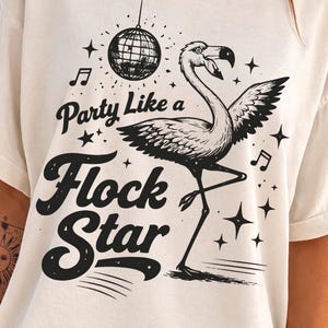 Puede incluir: Camiseta color crema con una ilustración negra de un flamenco, una bola de discoteca, notas musicales y estrellas. El texto dice "Party Like a Flock Star" en una fuente audaz y divertida.