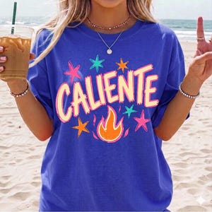 May include: A royal blue t-shirt with the word "CALIENTE" in a light orange font, surrounded by colorful stars and a flame graphic. The shirt is a casual style, perfect for warm weather.