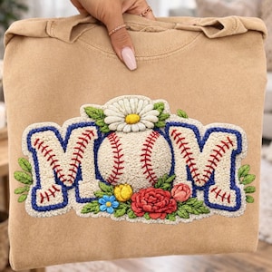 May include: Tan sweatshirt with a baseball-themed "MOM" design. The design features a baseball in place of the "O", with red stitching and a daisy on top. The letters are outlined in blue with red stitching.