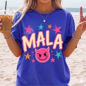 May include: A royal blue t-shirt with the word "MALA" in a light orange font, surrounded by pink, orange, and teal stars. Below the word is a pink devil face with a black outline. The shirt is a casual style.