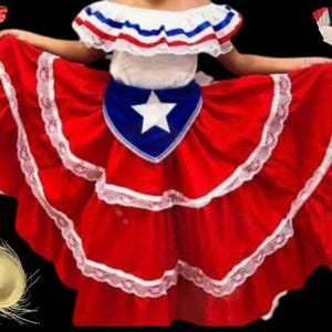 May include: A multi-tiered red skirt with white lace trim and a blue triangular panel featuring a white star. The top features an off-the-shoulder white ruffle neckline with red, white, and blue ribbon accents. Includes a straw hat and fan.