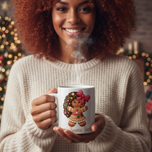 May include: A white ceramic mug featuring a gingerbread woman design with a pink bow and floral accents. The mug has the text "Sweetest Greetings" and is held by a person wearing a cream-colored sweater.