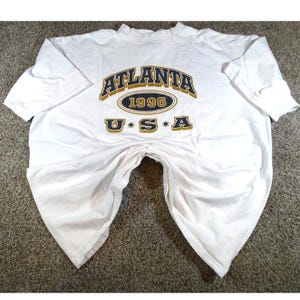 May include: White t-shirt with the text "ATLANTA 1996 USA" in gold and navy blue lettering. The shirt has short sleeves and a crew neck. The shirt is folded on a neutral-colored surface.