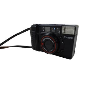 May include: A black Canon point-and-shoot film camera with a 38mm 1:2.8 lens, featuring an integrated flash, autofocus capabilities, and a black carrying strap attached to the side of the rectangular camera body.