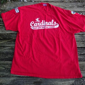 May include: A bright red short-sleeve t-shirt featuring the white text Cardinals and Cape Cod Baseball League with a small baseball player graphic. The shirt includes a rectangular patch on the right sleeve and a circular patch on the left sleeve.