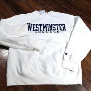 May include: A white Westminster College sweatshirt with long sleeves. The text "WESTMINSTER COLLEGE" is embroidered in navy blue across the chest. A small Champion logo is visible on the left sleeve cuff.