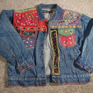 May include: A denim jacket with colorful embroidered patches. The jacket features red, green, and blue embroidery on the shoulders and pockets. The cuffs and hem are trimmed with red stitching. The jacket has a button-up front and a collar.
