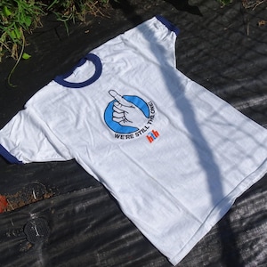 May include: A white ringer t-shirt with dark blue trim on the collar and sleeves. The chest features a graphic of a hand pointing upward inside a blue circle, with the text "WE'RE STILL THE ONE!" and a small red and blue "hth" logo below.