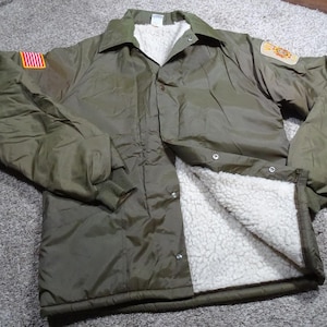 May include: Olive green jacket with a white, fluffy interior lining. The jacket has a collared neckline, button closures, and a patch with a flag design on the left sleeve. A second patch is on the right sleeve.