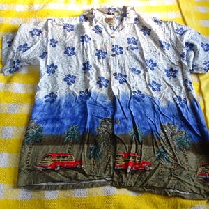 May include: A short-sleeved button-down shirt featuring a white upper section with a blue hibiscus floral pattern. The lower section displays a landscape print with palm trees and red vintage station wagon cars against a brown ground.