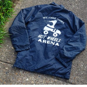 May include: A navy blue windbreaker jacket featuring a white graphic on the back. The graphic depicts a winged roller skate above the text MITE LEAGUE CHAMPS, HOT WHEELS ARENA. The jacket has long sleeves with elastic cuffs and a fold-down collar.
