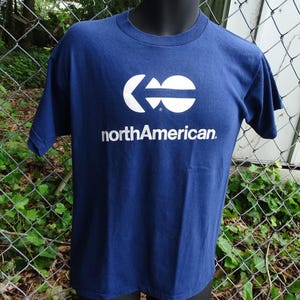 May include: A navy blue short-sleeved t-shirt featuring a white graphic logo on the chest. The logo consists of a stylized circular arrow icon above the text northAmerican printed in a white sans-serif font. The shirt has a crew neckline.