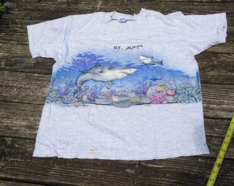 May include: A light gray t-shirt featuring a vibrant underwater scene with sharks, coral, and fish. The text "ST. JOHN" is printed above the image. The shirt is made of a soft material.