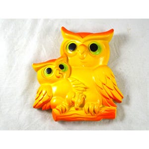 May include: A wall-mounted decorative plaque featuring a large yellow owl and a smaller yellow owl perched together on a branch. Both owls have bright orange accents on their ears and wings, with large black eyes and green highlights.