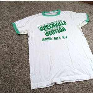May include: White t-shirt with green trim around the collar and sleeves. The shirt features the text "GREENVILLE SECTION" in green, with "JERSEY CITY, N.J." printed below.
