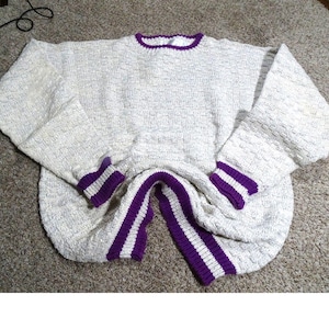 May include: Cream-colored sweater with purple and white striped accents on the cuffs and lower hem. The sweater has a crew neck with a purple trim. The sleeves are long and the material appears to be a textured knit.