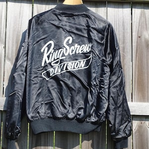 May include: Black satin bomber jacket with the text "Ring Screw DIVISION" in white script across the back. The jacket has a black ribbed collar, cuffs, and hem. The jacket is hanging on a wooden hanger.