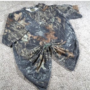 May include: A camouflage-patterned t-shirt with a pocket on the chest. The shirt features a blend of brown, green, and black hues, mimicking a forest environment. The short-sleeved shirt is made of a soft material and is suitable for outdoor activities.