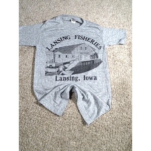 May include: A gray t-shirt featuring a black and white graphic of a building with the text "LANSING FISHERIES" above and "Lansing, Iowa" below. The shirt is made of a soft material and has short sleeves.
