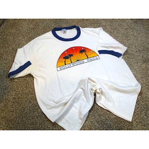 May include: White t-shirt with a navy blue collar and sleeve trim. The front features a graphic of palm trees, a sunset, and the text "HOLLYWOOD YMCA". The shirt is made of a soft, lightweight material.