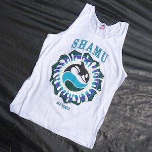 May include: A white sleeveless tank top featuring a graphic print of an orca whale surrounded by a blue and green splash design. The text SHAMU is printed in blue above the graphic, and the text SEA WORLD is printed in blue below the graphic.
