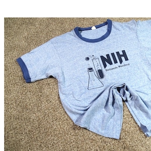 May include: Light blue ringer t-shirt with navy blue trim. The shirt features a graphic of laboratory equipment and the text "NIH Bethesda, Maryland" in navy blue.