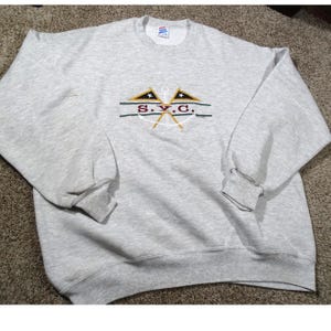 May include: A heather gray sweatshirt with a crew neck and long sleeves. The front features an embroidered design with two flags, anchors, and the letters "S.Y.C." in a horizontal line. The cuffs and hem are ribbed.