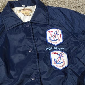 May include: A navy blue Antigua jacket with a white collar and two patches. The patches feature an anchor design with the text "PWBA" and "High Hampton" embroidered in blue.