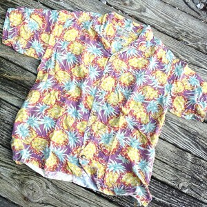 May include: A short-sleeve button-down shirt featuring an all-over pineapple print in shades of yellow, light blue, and purple. The shirt has a classic collar, a front button placket, and a single chest pocket. The fabric displays a repeating pattern.