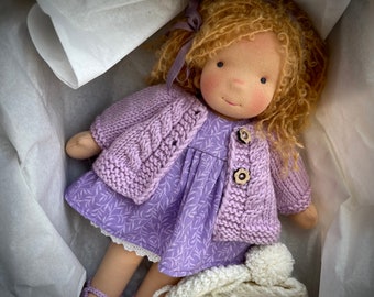 May include: A handmade doll with curly blonde hair, wearing a lavender dress, cable-knit cardigan, and matching shoes. The doll has a sweet, painted face and is nestled in a white box with a knitted hat.