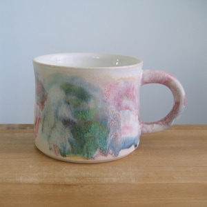 Op de afbeelding: Een keramische mok met een witte binnenkant en een kleurrijke buitenkant. De buitenkant van de mok heeft een aquarelontwerp met roze, groene, blauwe en paarse tinten. De mok heeft een afgerond oor en staat op een houten oppervlak.