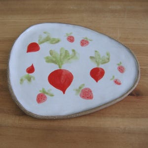 May include: A white, irregularly shaped ceramic plate with a hand-painted design of red radishes and strawberries with green leaves. The plate has a textured, light brown rim and is set on a wooden surface.