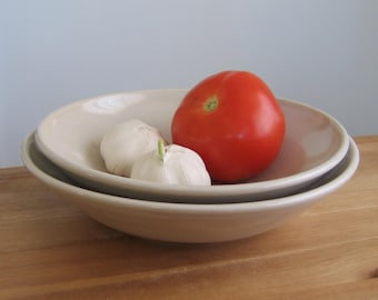 May include: Two white garlic cloves and a red tomato in a beige ceramic bowl. The bowl is nested inside a larger beige ceramic bowl.