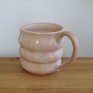 May include: A handmade ceramic mug with a soft, pale pink glaze and a distinctive ribbed, undulating body shape. The mug features a smooth, glossy finish and a curved handle attached to the side, showcasing a subtle variation in color intensity.