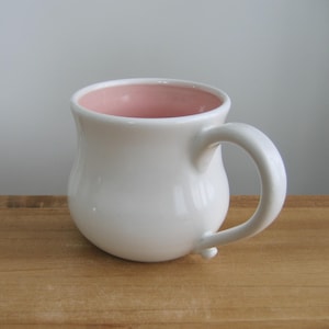May include: A white ceramic mug with a rounded body and a curved handle. The interior of the mug is a soft pink color. The mug is sitting on a wooden surface.