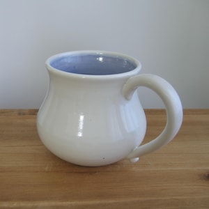 May include: A white ceramic mug with a rounded body and a curved handle. The interior of the mug is light blue. The mug is sitting on a wooden surface.