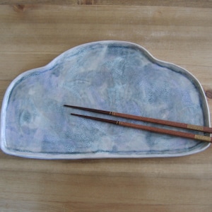 Può includere: Un piatto da portata in ceramica a forma di nuvola con smalto marmorizzato blu e bianco. Due bacchette marroni con dettagli dorati poggiano sul piatto. Il piatto è su una superficie di legno.
