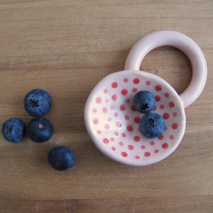 May include: A pink ceramic dish with red polka dots and a raised ring handle. The dish is filled with blueberries.
